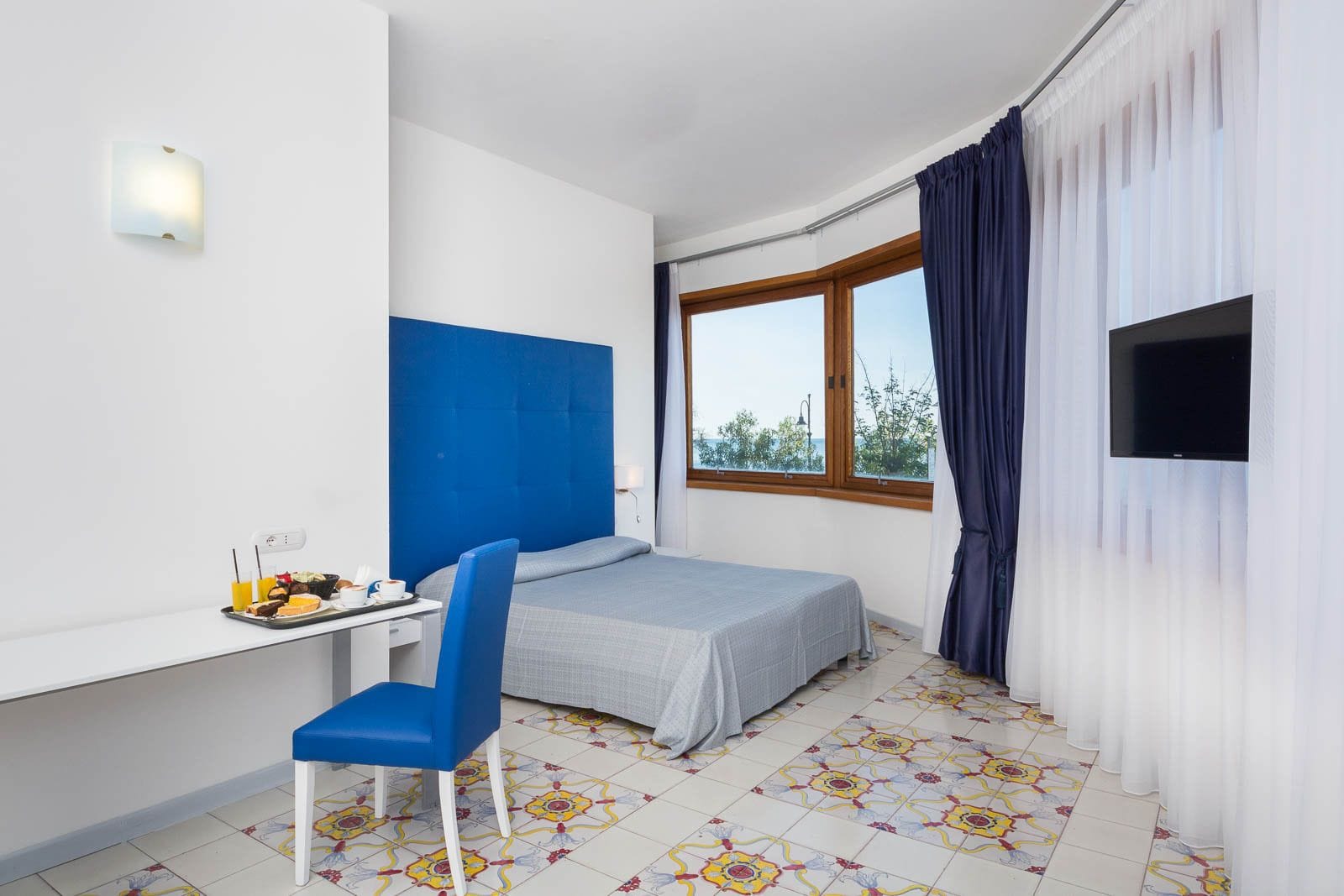 A hotel room with a bed, a desk with a chair, a TV, and a window with curtains.