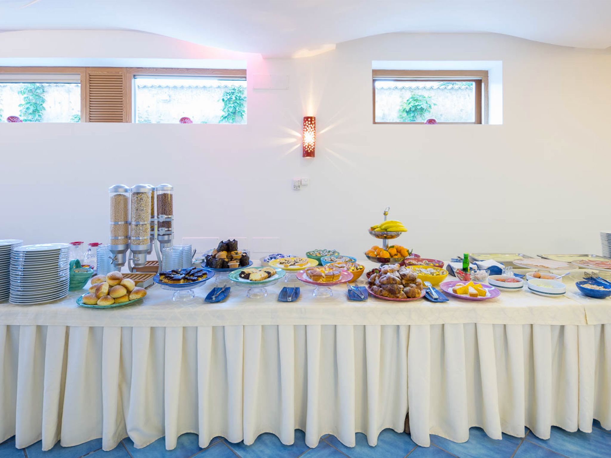 Una mesa de buffet con diversos alimentos y aderezos, ubicada en una sala con ventanas y un accesorio de iluminación en la pared.