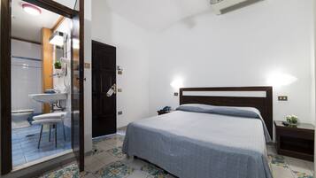 A bedroom with a bed, a bedside table, a nightstand, a small table with a plant, and a bathroom visible through an open door.
