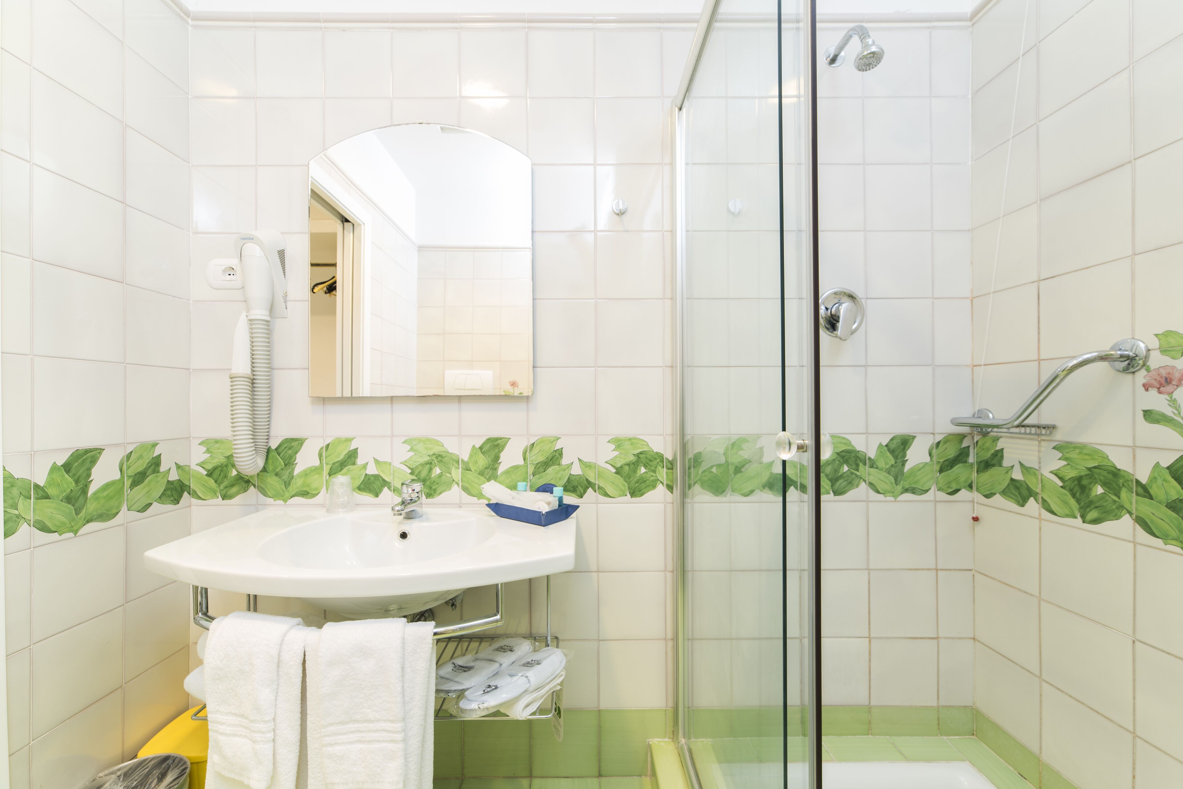 A bathroom with a white sink, a glass shower enclosure, and green leaf-patterned tiles.