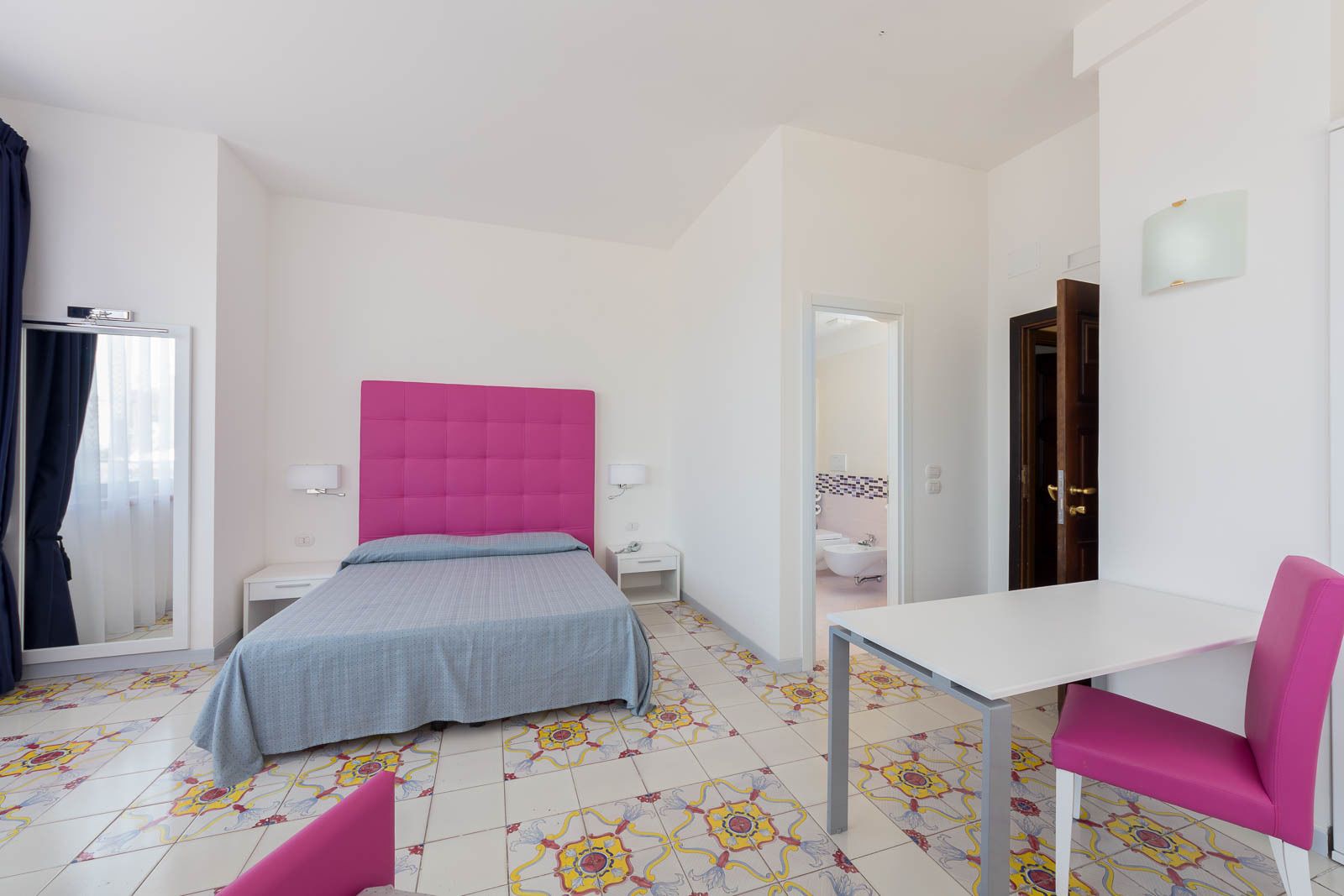 A hotel room with a bed, a desk, a chair, a bathroom, and a mirror.