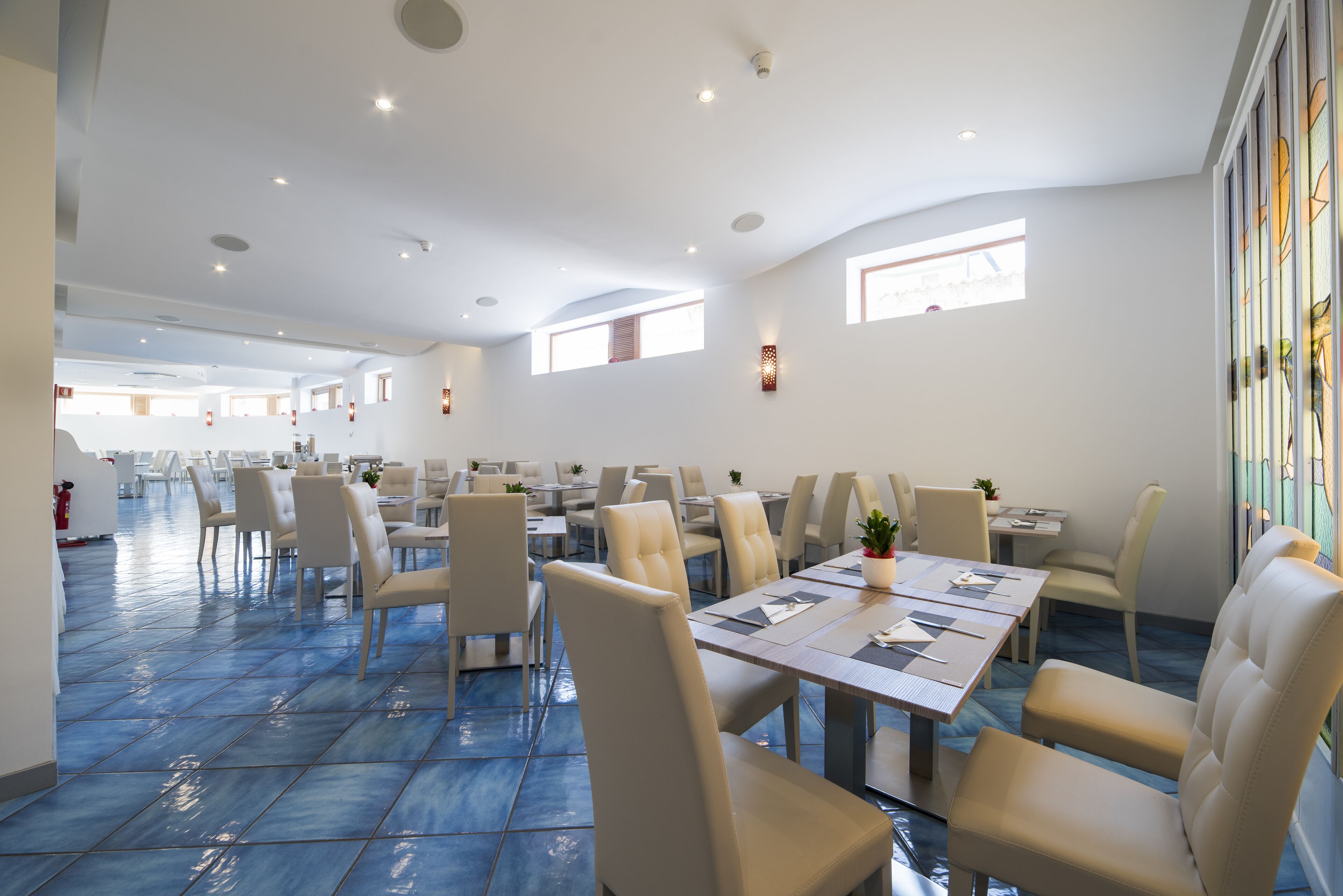 Un vaste espace dédié à la salle à manger, avec de nombreuses tables et chaises, de grandes fenêtres et un sol recouvert de carreaux bleus.