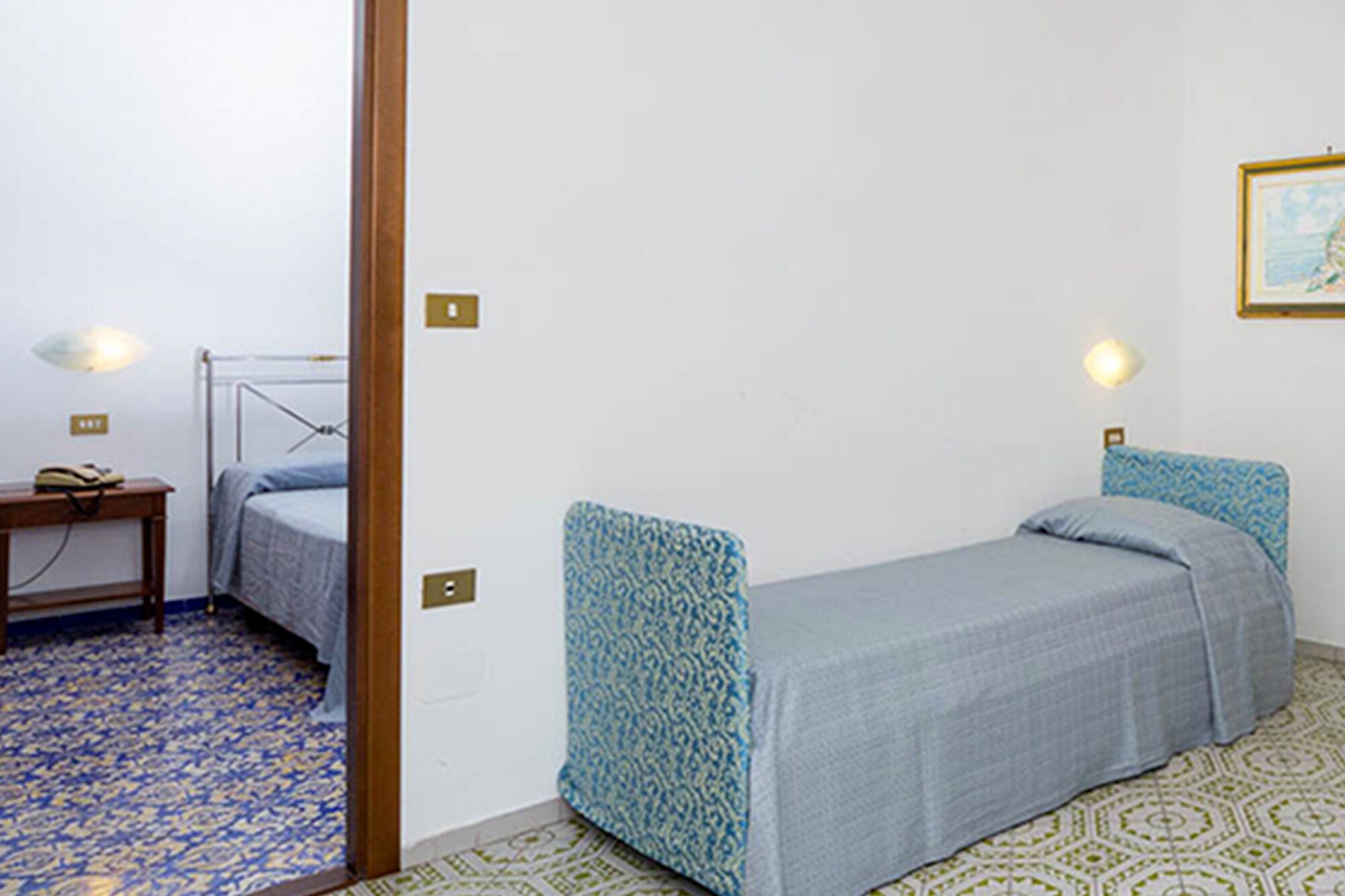 A small, single-bed room with patterned bedding, a bedside table, and wall-mounted lights.