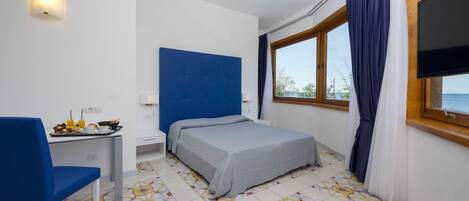 Una habitación con cama, escritorio con desayuno, televisor y ventana con cortinas.
