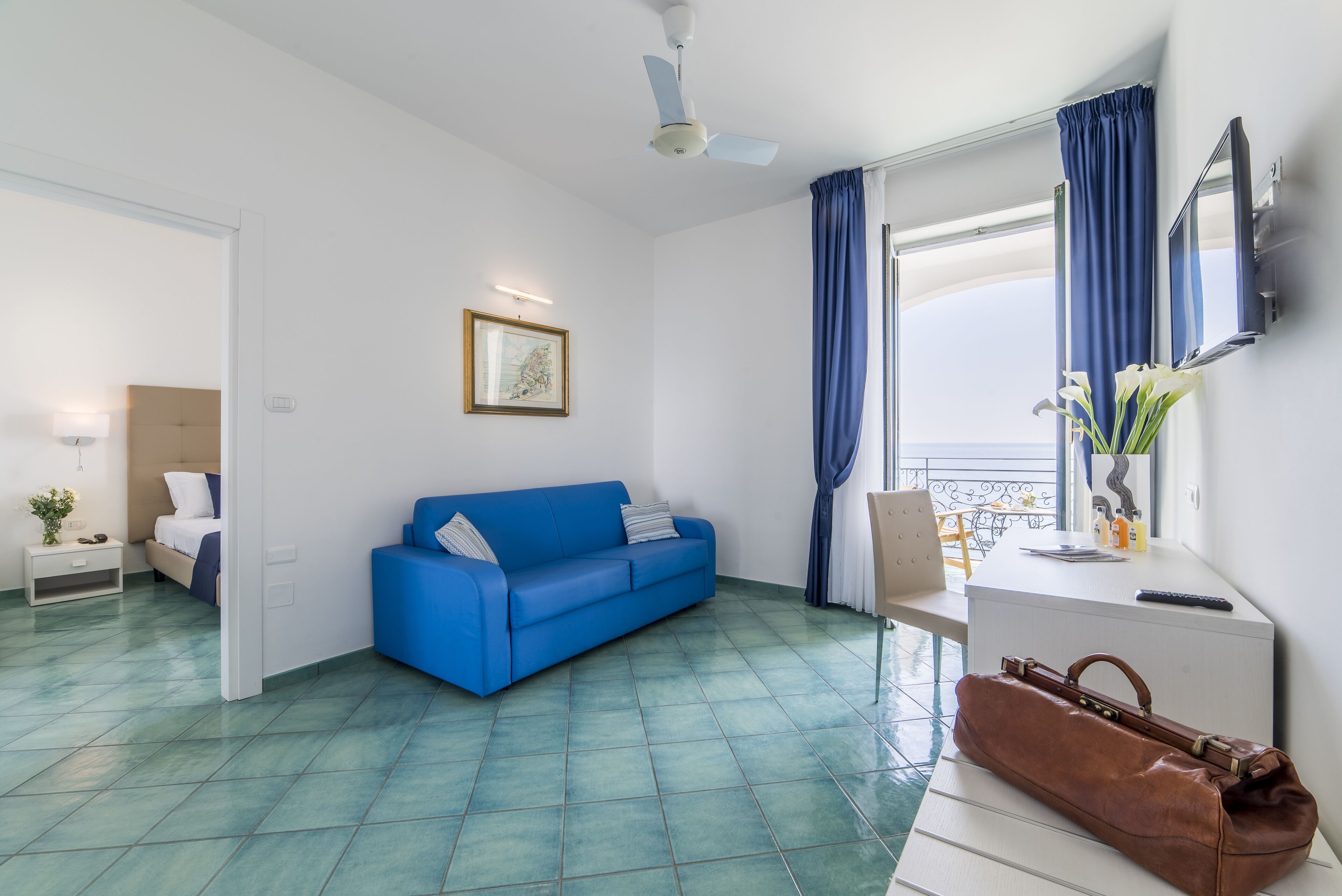 Una habitación moderna con un sofá azul, un escritorio blanco, un bolso de cuero marrón y un ventilador de techo.