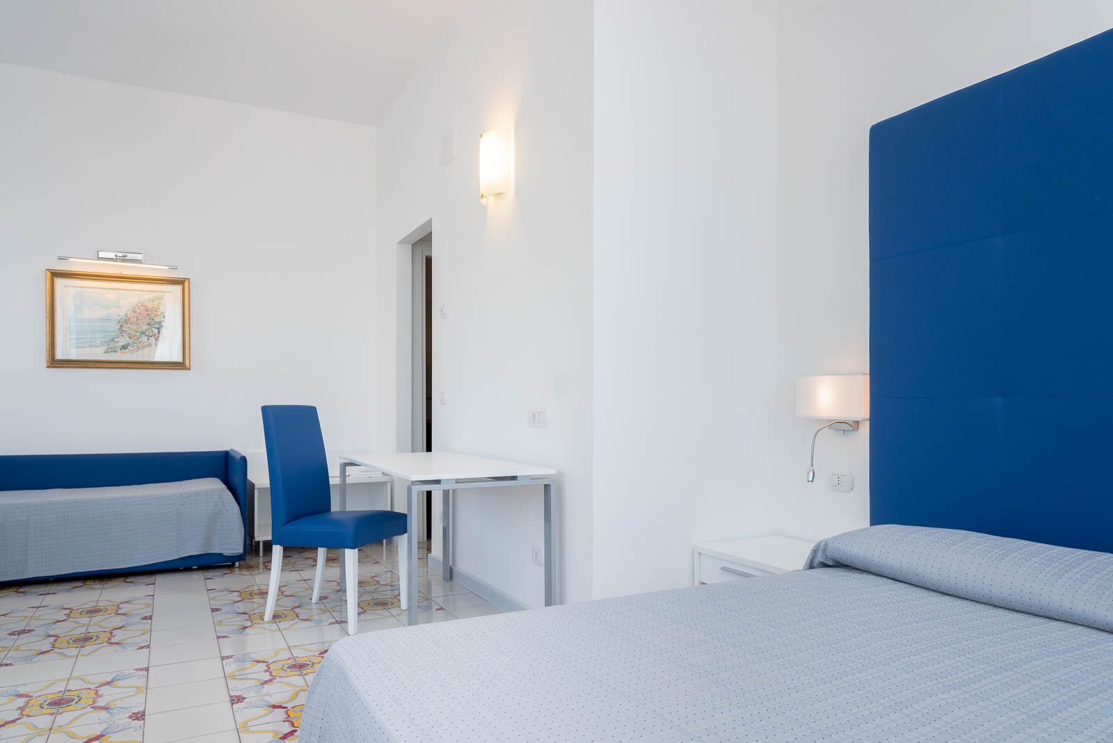 A hotel room with two beds, a desk, a chair, a small table, and a wall-mounted lamp.