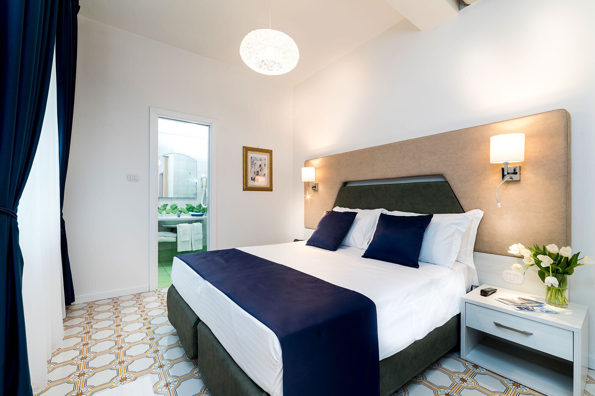 A hotel room with a bed, bedside table, nightstand, and a view of the outside.