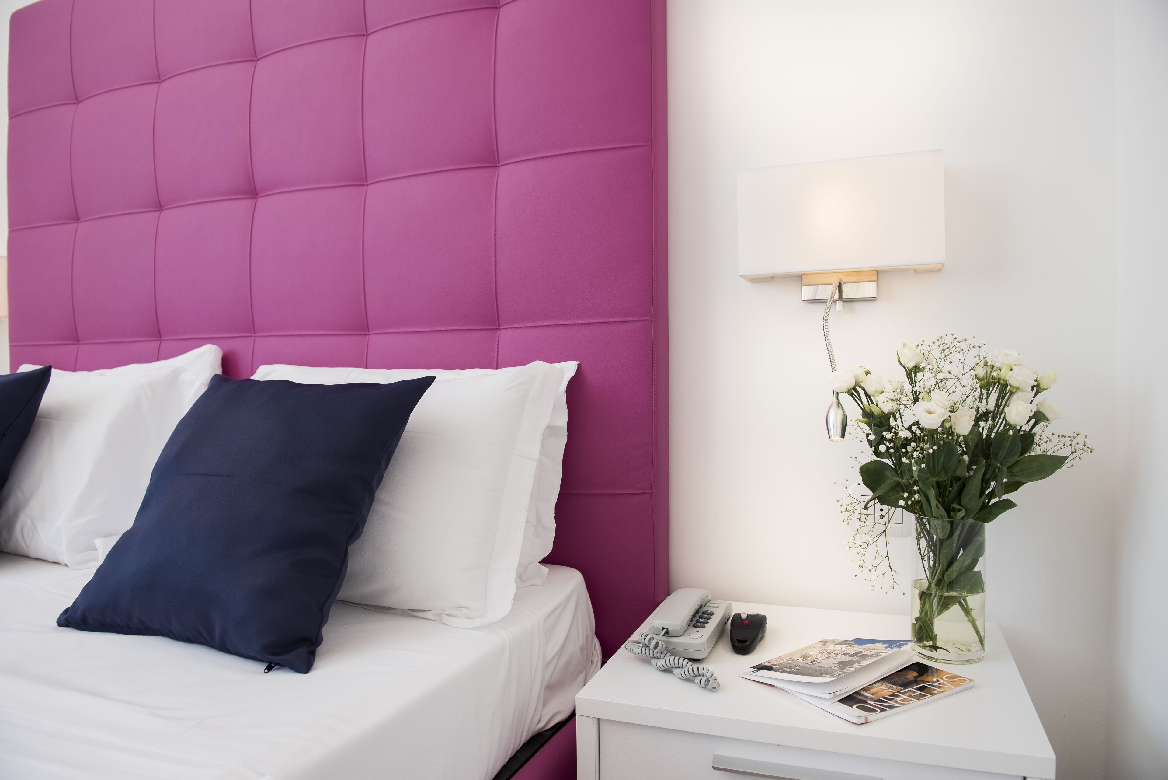 Habitación de hotel con una cama, ropa de cama blanca, cabecera rosa, mesita de noche con un teléfono, un jarrón con flores y una lámpara de pared.