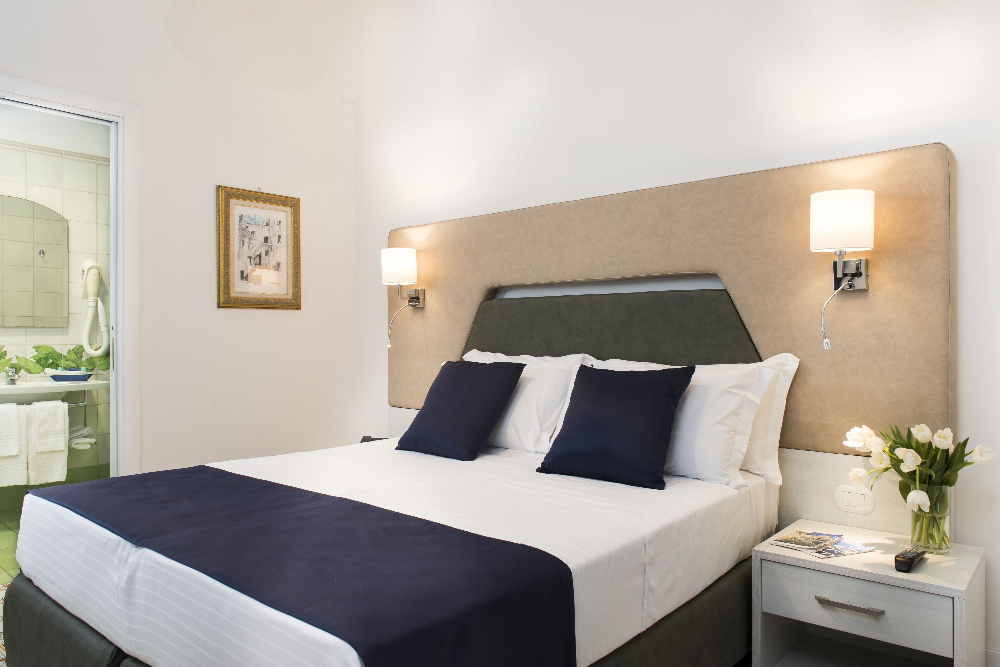 A neatly made bed with a dark blue bedspread and white linens, a bedside table with a lamp, and a framed picture on the wall.