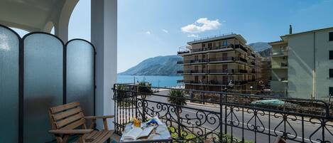 A balcony with wooden chairs, a table with a book and a glass, and a view of a coastal building and sea.