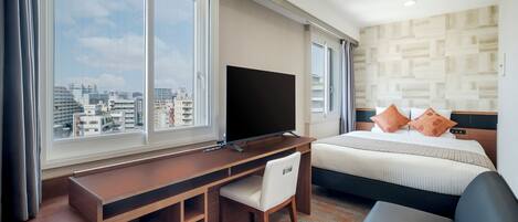 A hotel room with a bed, a desk with a chair, a television, and large windows offering a city view.