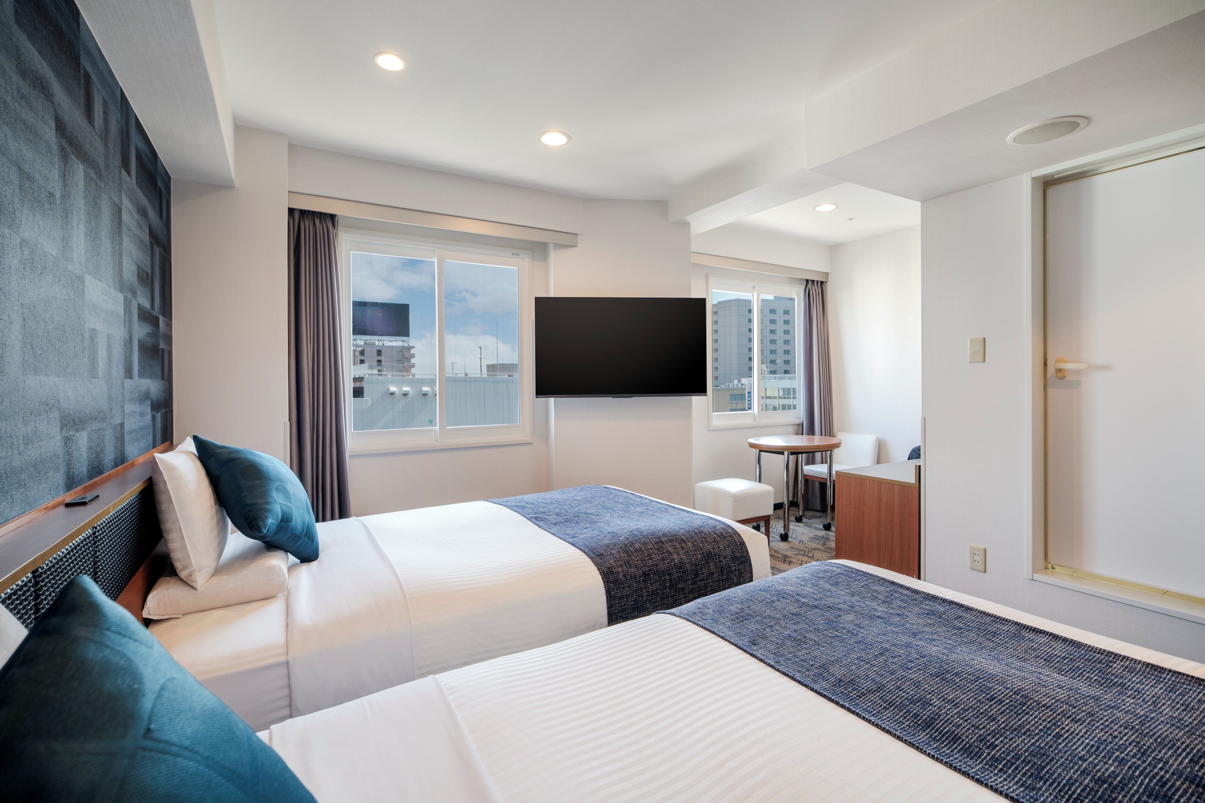 A hotel room with two beds, a TV, a desk, and a view of the city.