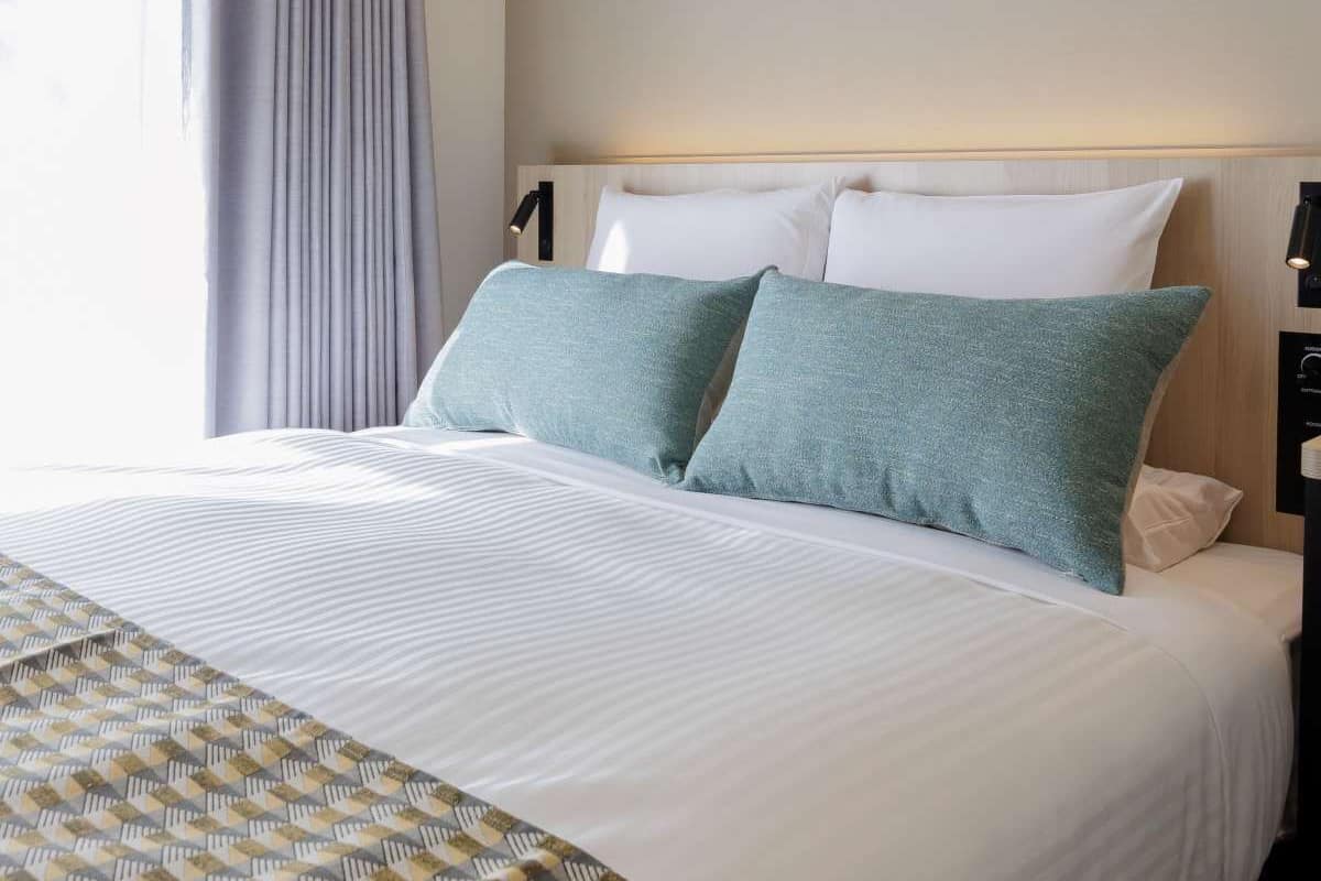 Una cama bien hecha con ropa blanca y dos almohadas color turquesa, un cabecero de madera y una lámpara de noche.