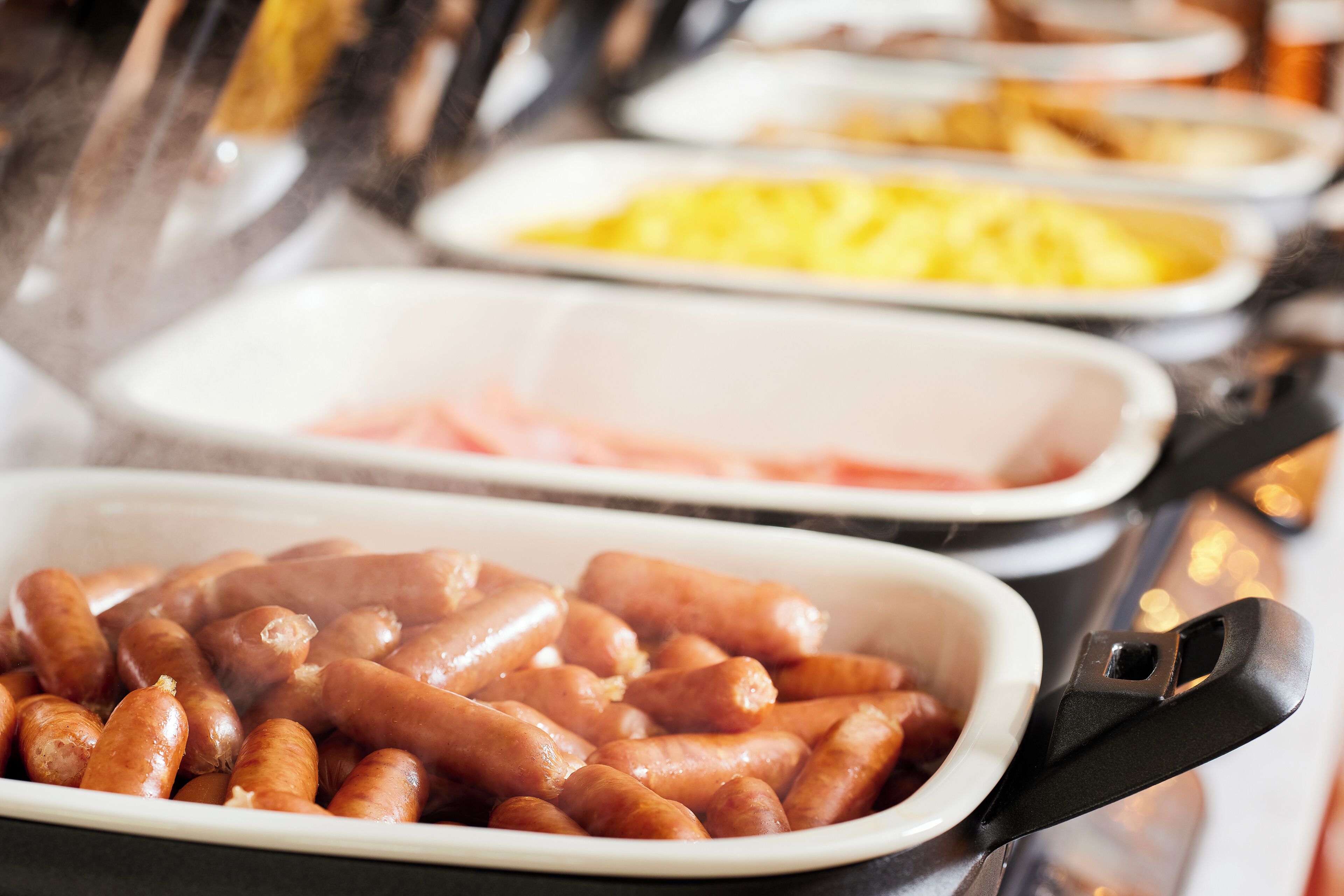 A buffet with various food items including sausages, ham, and scrambled eggs.