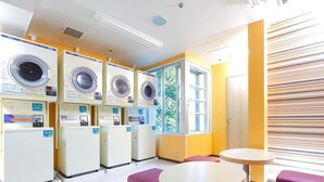 Une buanderie équipée de plusieurs machines à laver, une fenêtre donnant sur un espace verdoyant et un coin salon avec une table ronde et deux chaises.