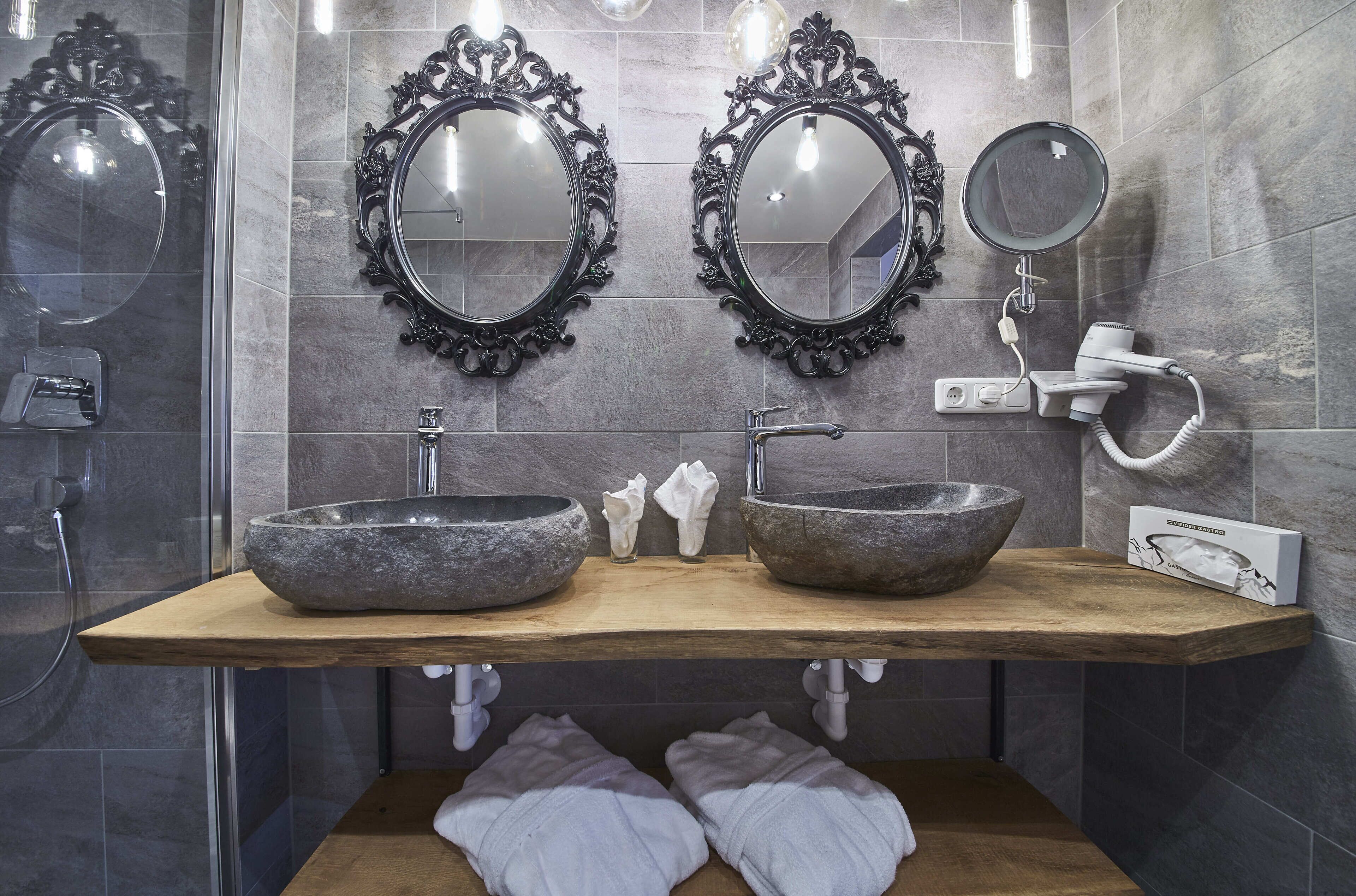 Een badkamer met twee stenen wasbakken, sierlijke spiegels, een handspiegel en een haardroger.