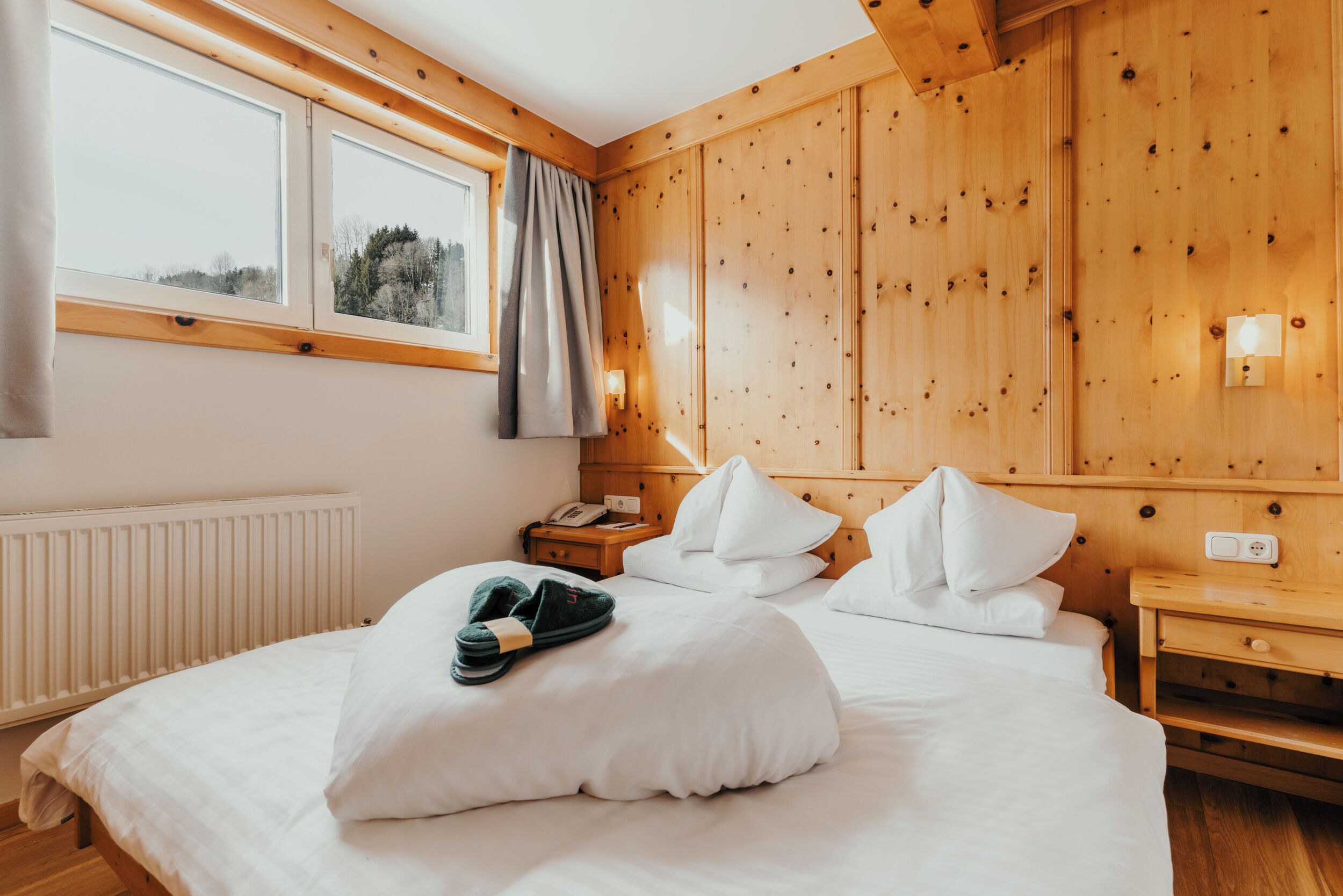 Een kamer met houten lambrisering, een bed, nachtkastje, raam met gordijnen en een wandlamp.