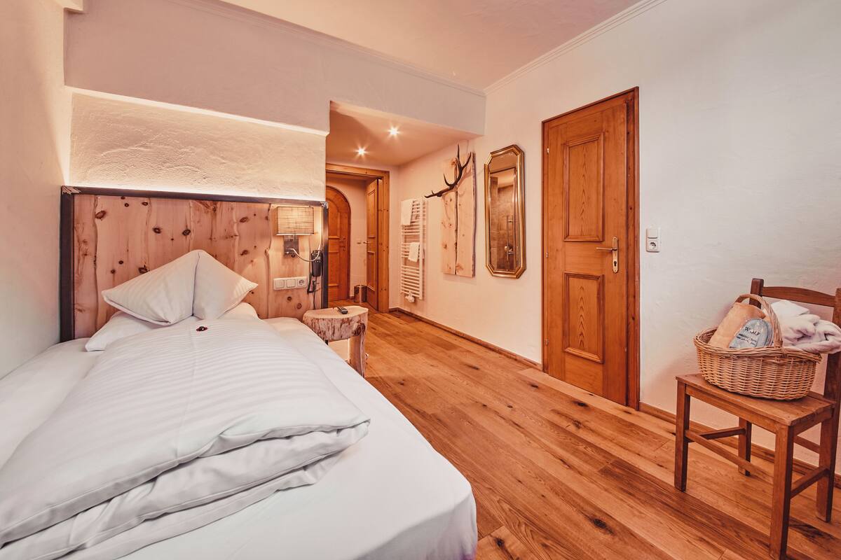 Un dormitorio con cabecera de madera, cama con ropa de cama blanca, suelo de madera, puerta de madera y una silla de mimbre con cesta.