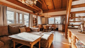 Ein gemütliches Zimmer mit Holztisch, Sofa und Blick auf die Berge durch große Fenster.