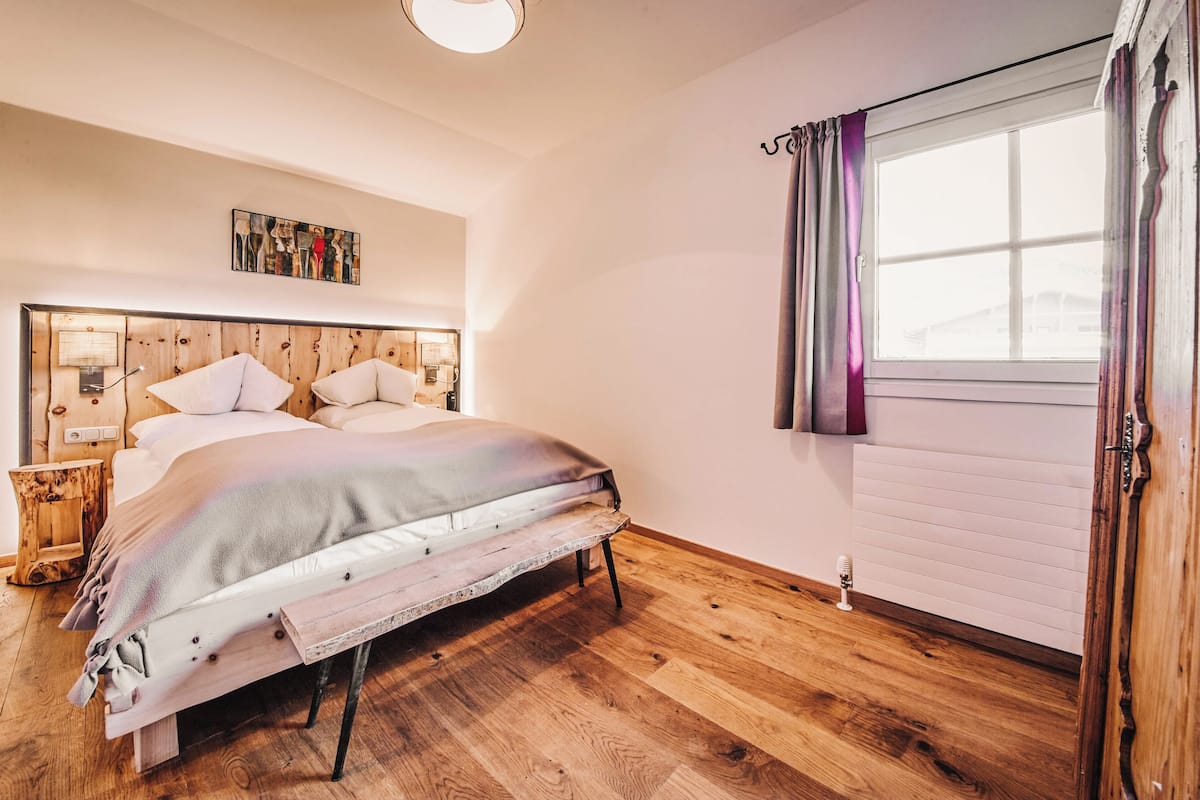 Un dormitorio con cabecera de madera, una cama con ropa de cama blanca, una mesita de noche con una lámpara y una ventana con cortinas.