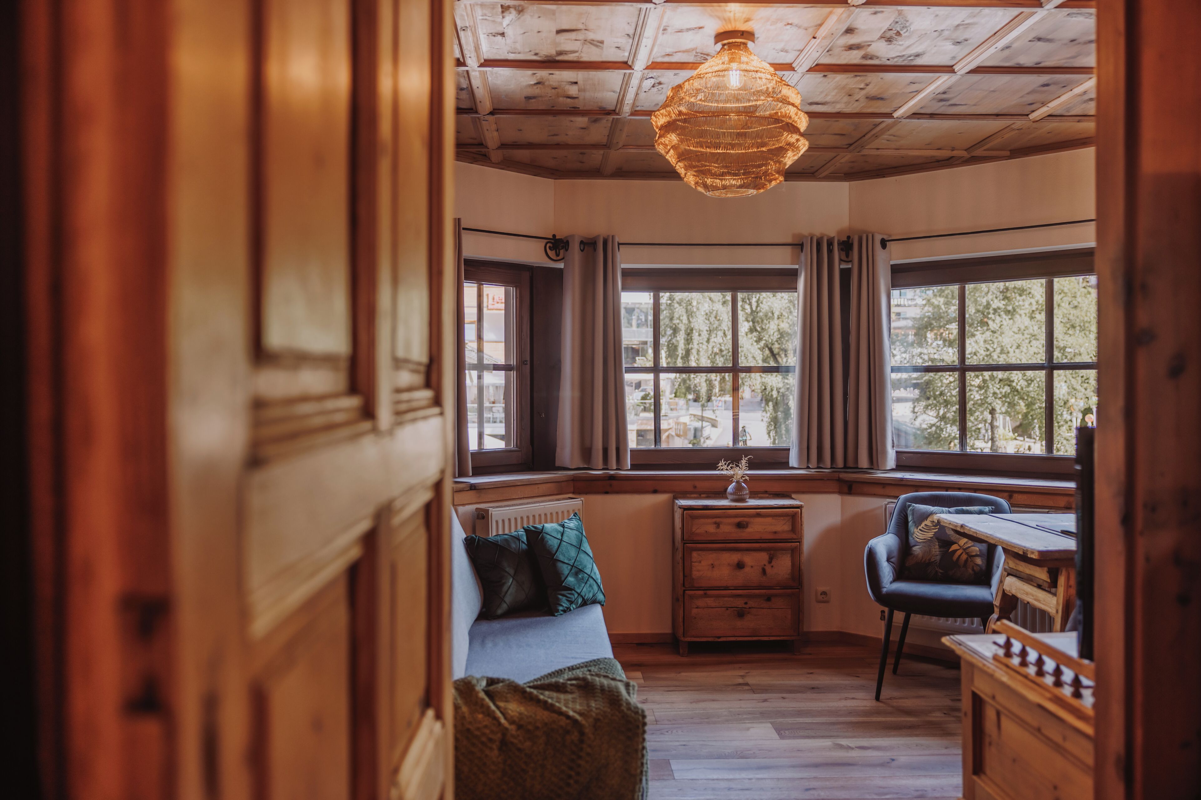 A cozy room with a wooden ceiling, a large window with curtains, a wooden dresser, a chair, and a small table.