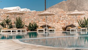 A pool area with lounge chairs and umbrellas, surrounded by a stone wall and a mountain backdrop. - Margarenia (Santorini)