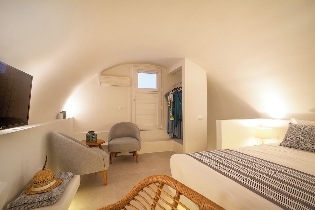 A bedroom with a bed, a chair, a small table, a TV, and a window.