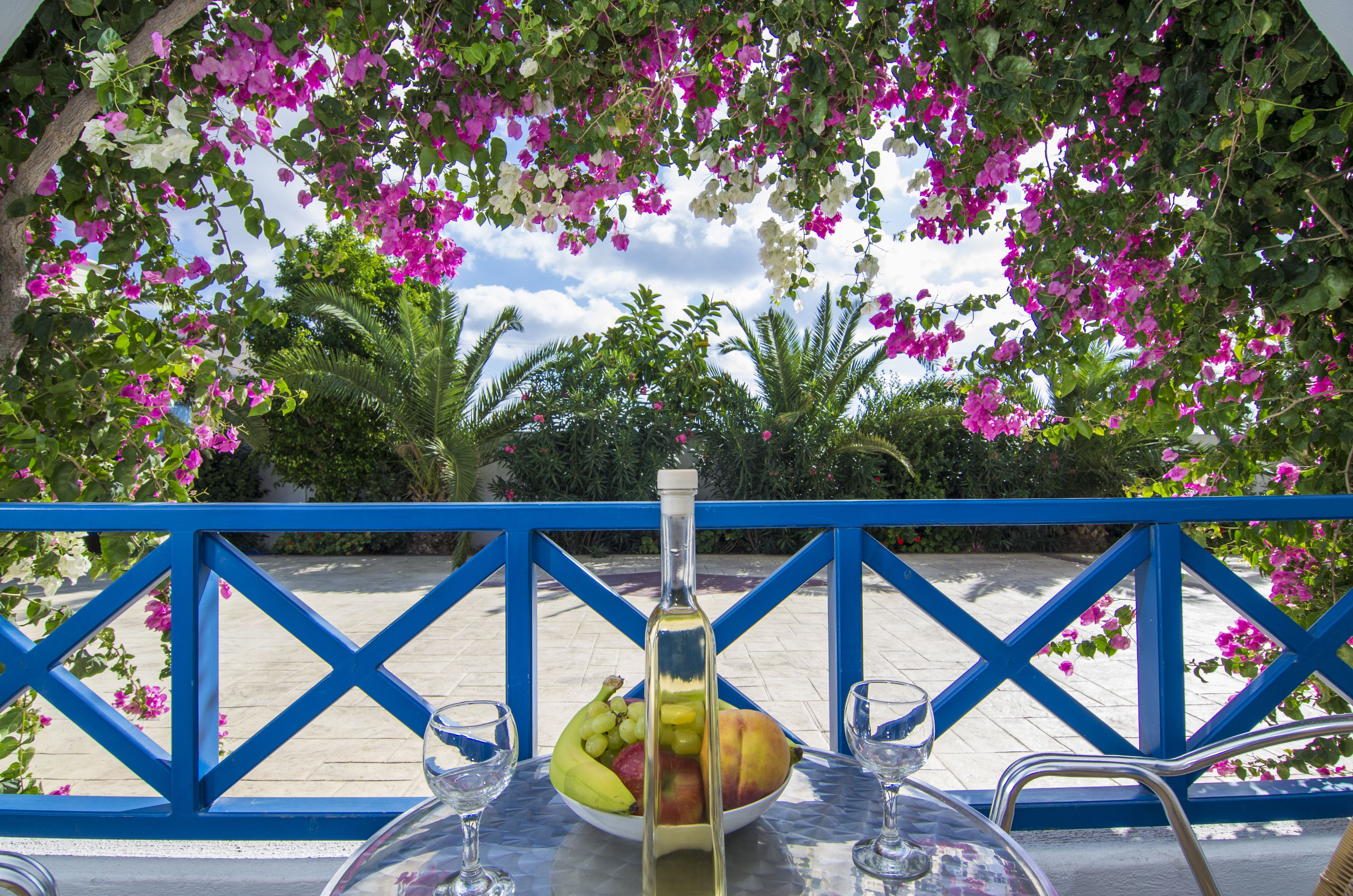 A table with a bottle, a bowl of fruit, and two glasses, set against a blue railing with pink flowers in the background.