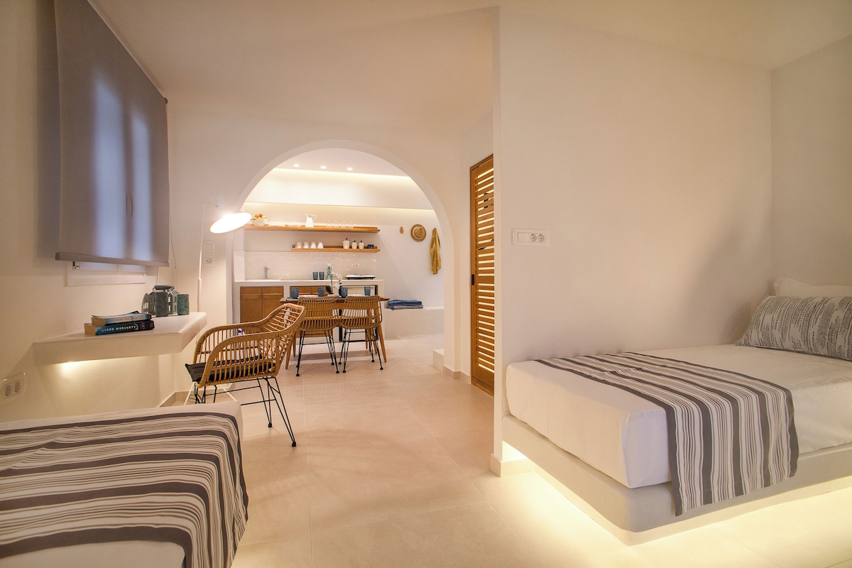 Una habitación de hotel moderna con una cama, un pequeño comedor con sillas y una mesa, y una cocina al fondo.