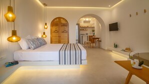 A modern hotel room with a large bed, bedside lamps, a small table with a vase, and an arched doorway leading to a kitchenette. - Margarenia (Santorini)