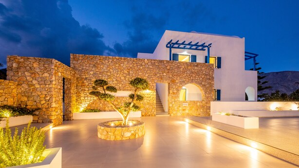 A modern building with stone walls and a flat roof, featuring a well-lit outdoor area with planters and a staircase. - Margarenia (Santorini)