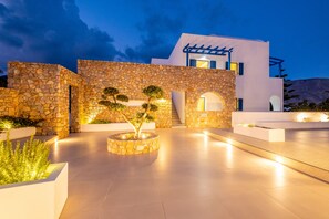 A modern building with stone walls and a flat roof, featuring a well-lit outdoor area with planters and a staircase. - Margarenia (Santorini)