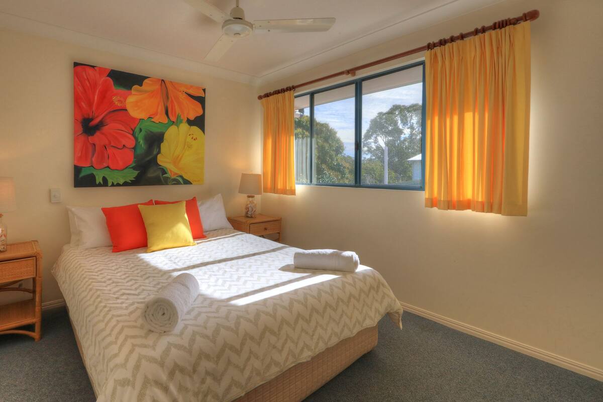 A bedroom with a bed, a nightstand, a lamp, a painting, and a window with curtains.