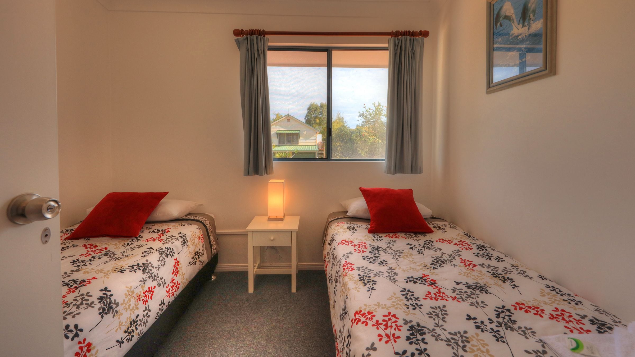 A bedroom with two single beds, a nightstand with a lamp, a window with curtains, and a painting on the wall.