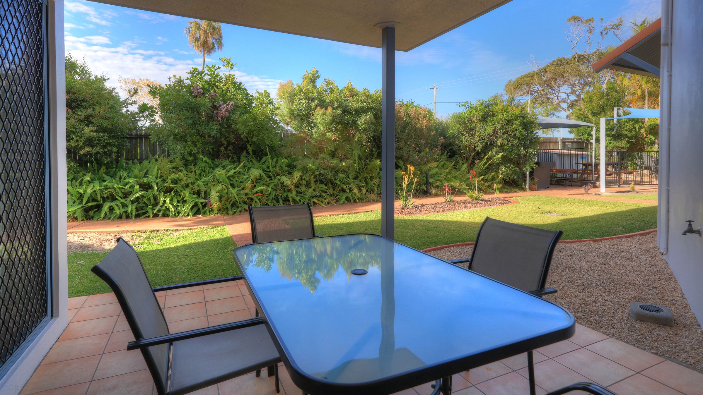 Une terrasse extérieure avec une table et des chaises en verre, entourée d’un jardin où poussent différentes plantes et arbres.