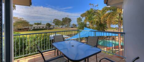 A balcony with a table and chairs, overlooking a swimming pool and a grassy area with trees.