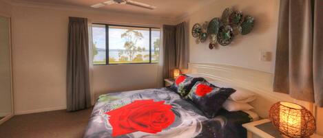 A bedroom with a bed, a ceiling fan, a window with curtains, a bedside table with a lamp, and wall art.