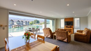 A spacious living room with a large window overlooking a pool and mountains, a wooden dining table, and comfortable seating. - Lake Resort by Marsden (Mount Pisa)