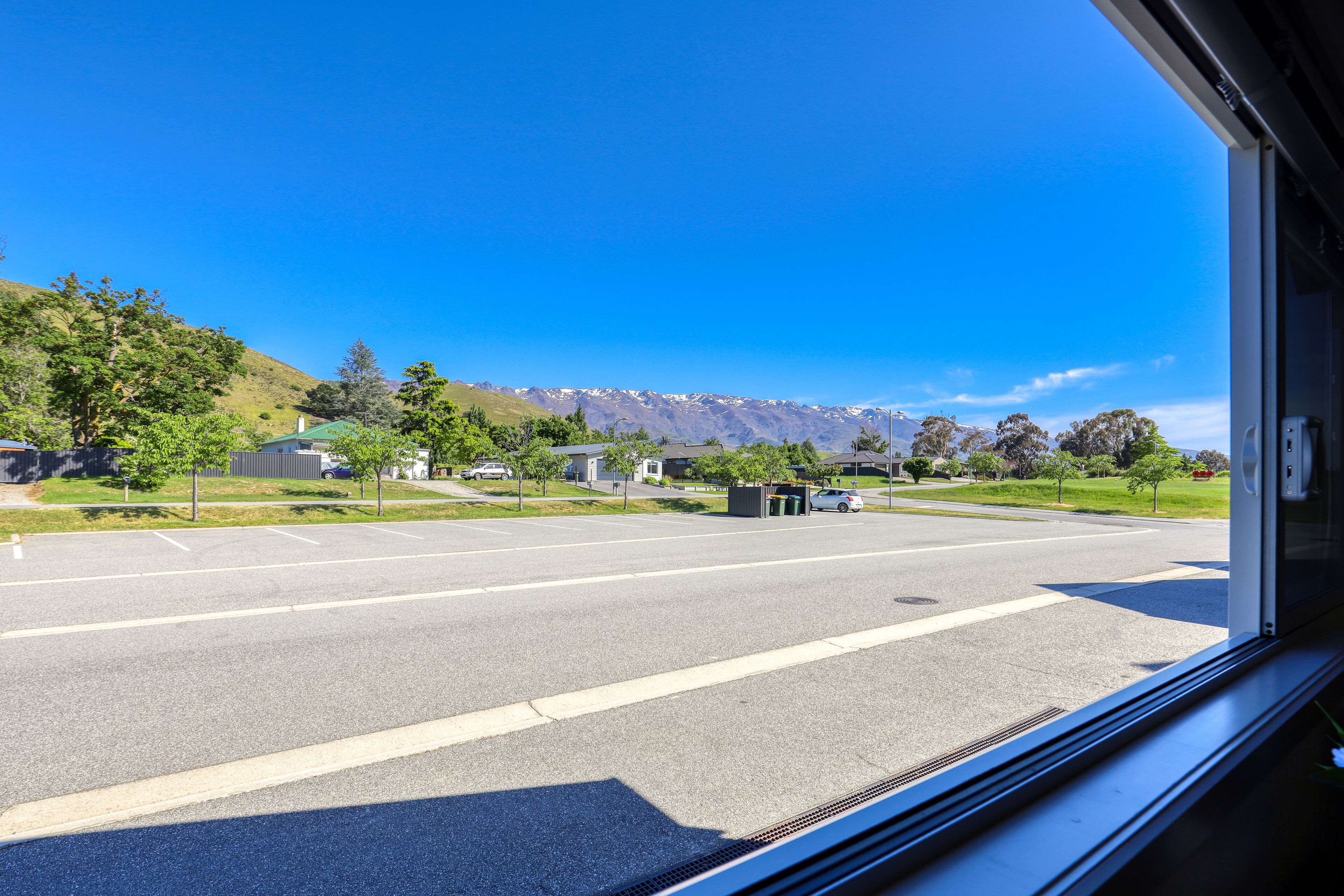 A view from inside a vehicle, looking out onto a street with houses, a parking lot, and mountains in the background.