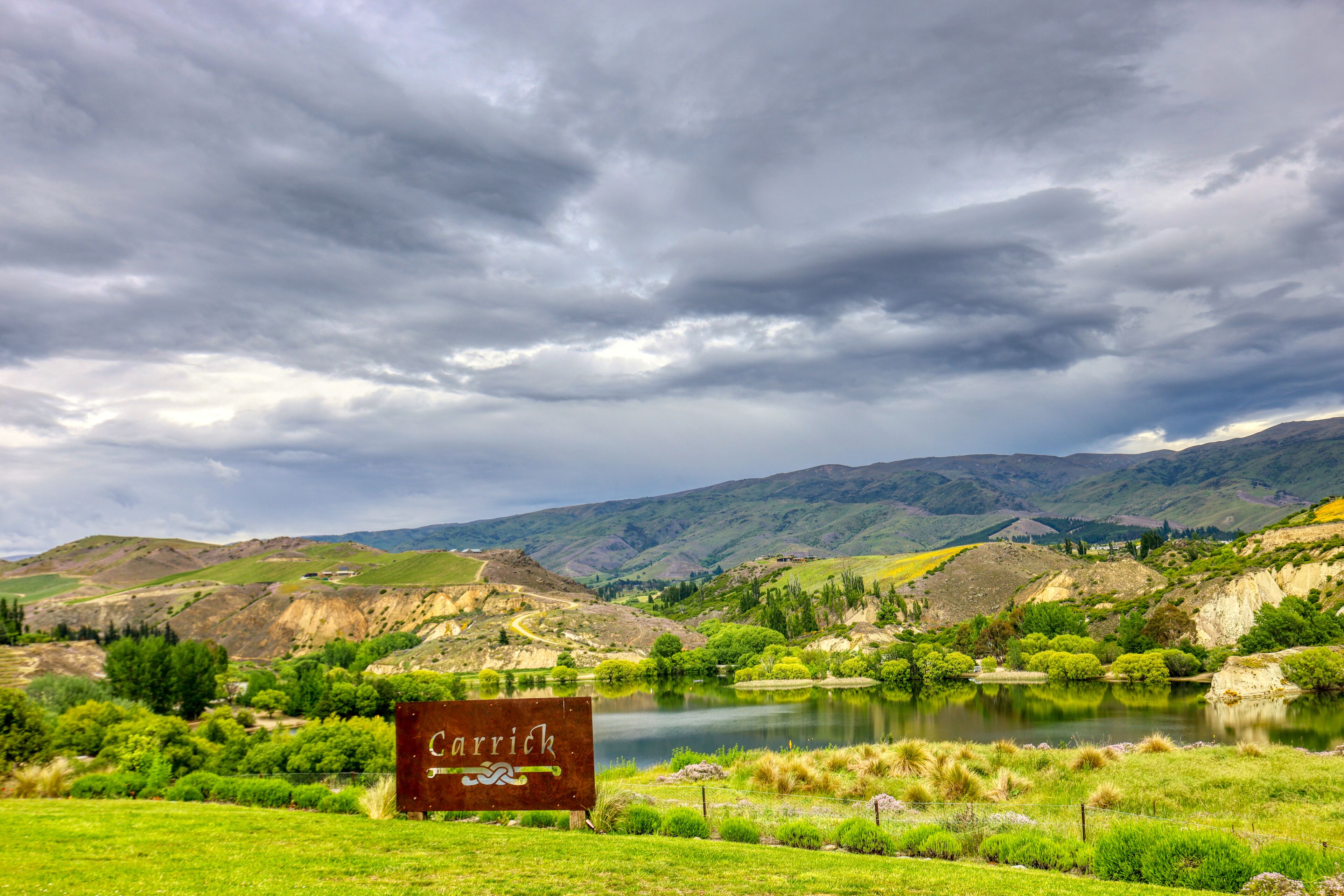 A sign reading "Carrick" in a rural landscape with a lake, hills, and a cloudy sky.