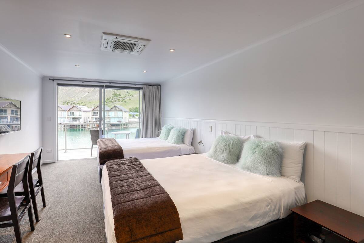 A hotel room with two beds, a desk, and a view of a water body and buildings.