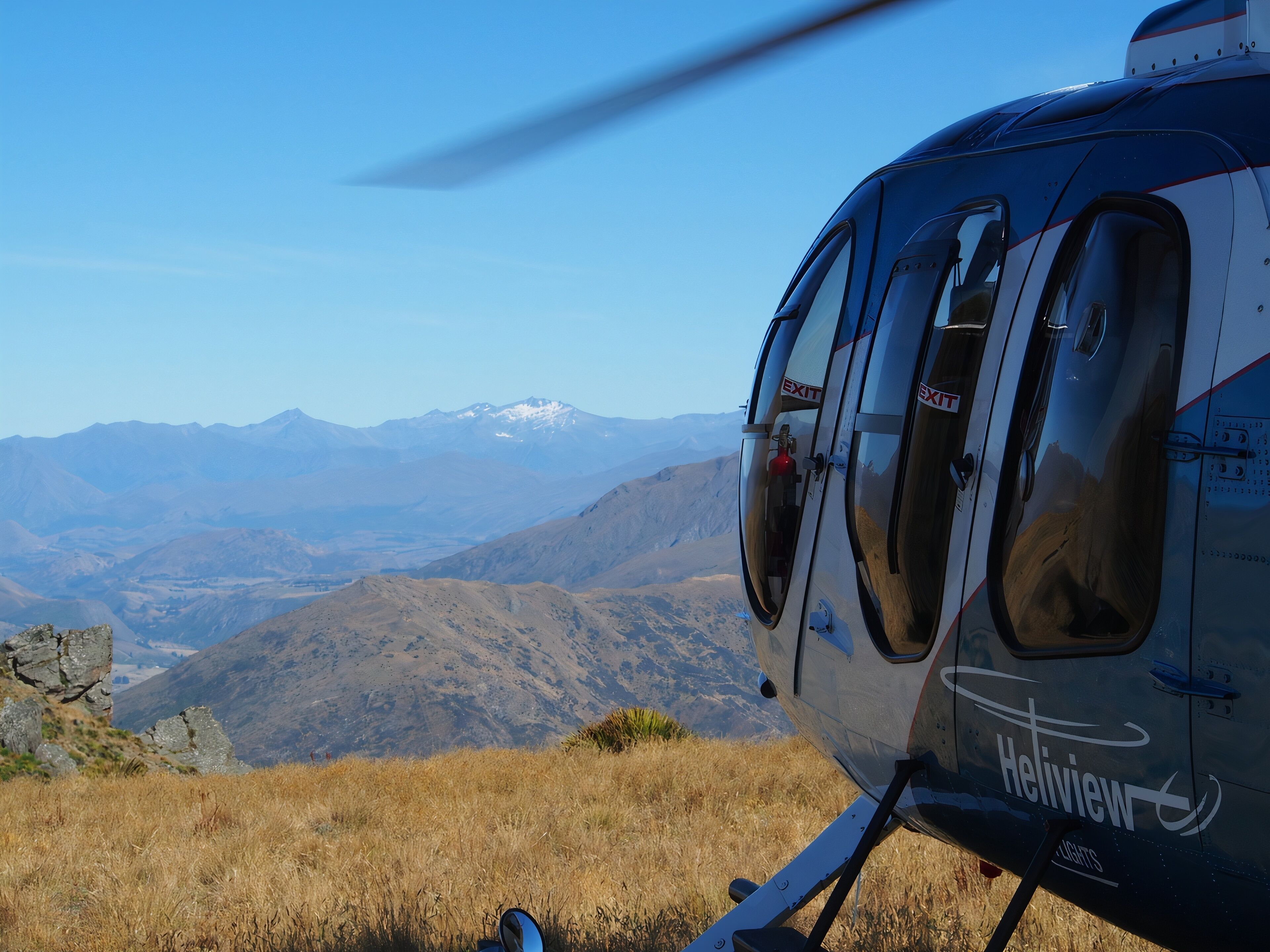 Een helikopter met de branding 'Helitview' geparkeerd op een grasheuvel, met een bergachtig landschap op de achtergrond.