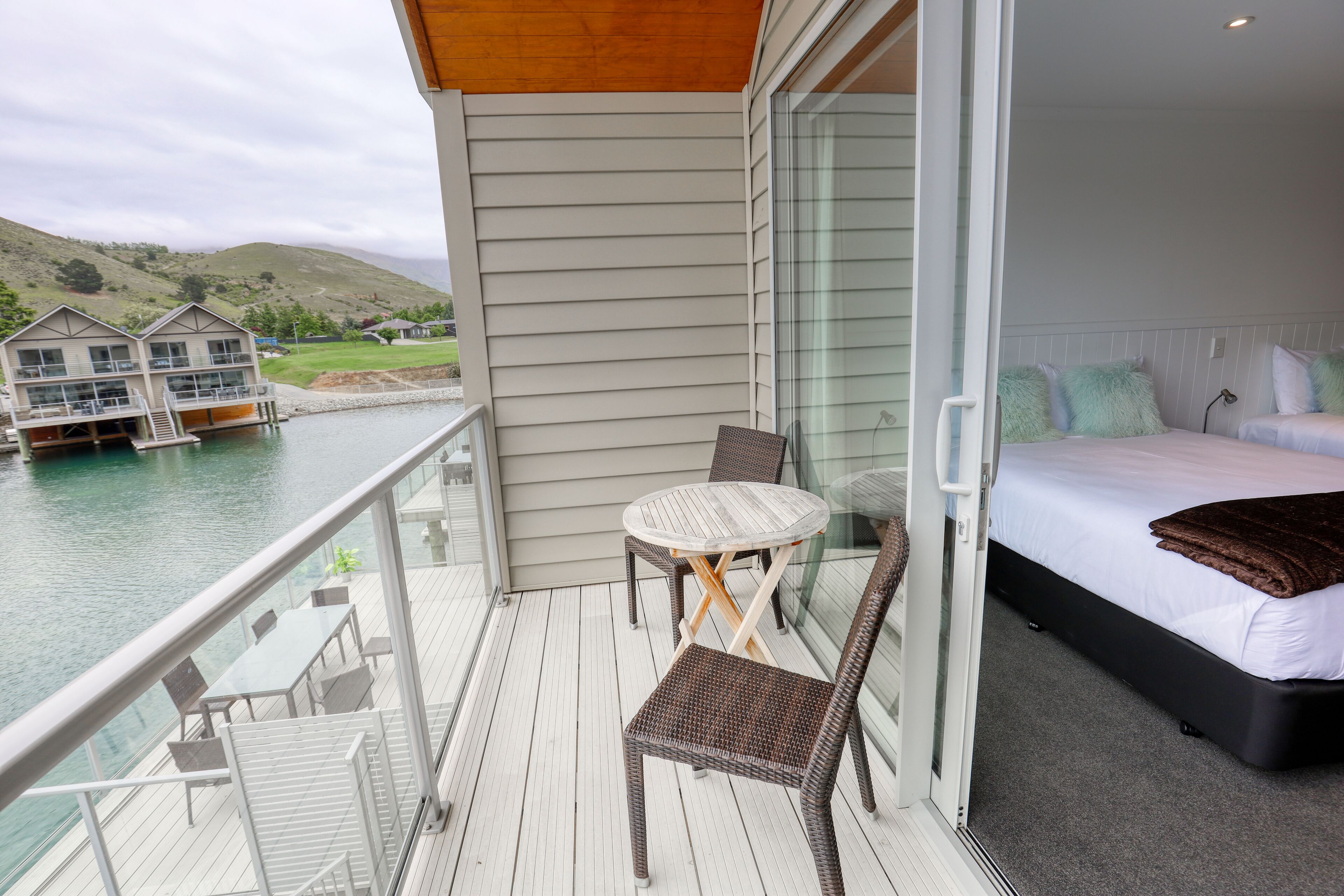 A balcony with a table and chairs, a bed with pillows, and a view of a water body and buildings.