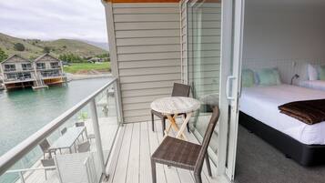 A balcony with a table and chairs, a bed with pillows, and a view of a water body and buildings.