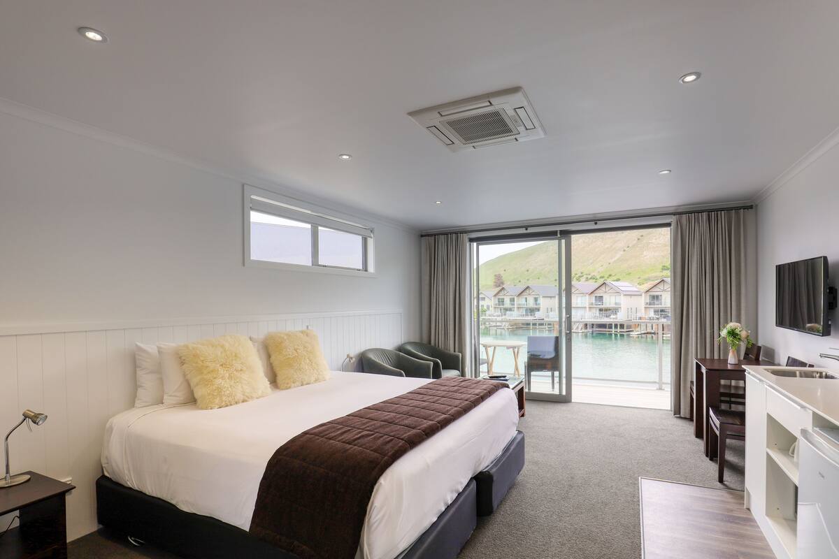 A hotel room with a large bed, a desk, a chair, a TV, and a view of a body of water and buildings.
