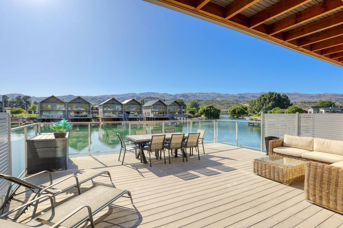 A wooden deck with a table and chairs, overlooking a body of water and mountains.