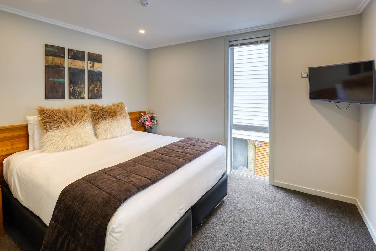 A bedroom with a bed, a flat-screen TV, and a window with blinds.