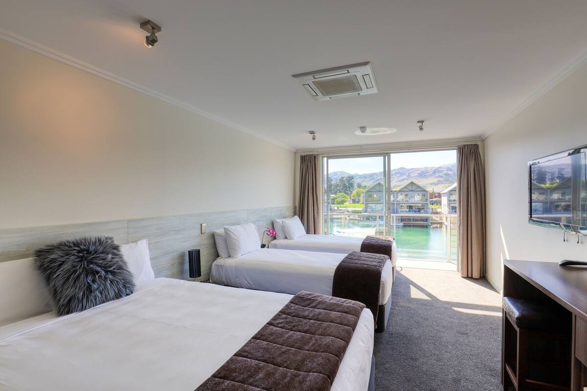 A hotel room with three beds, a TV, and a view of a pool and buildings.