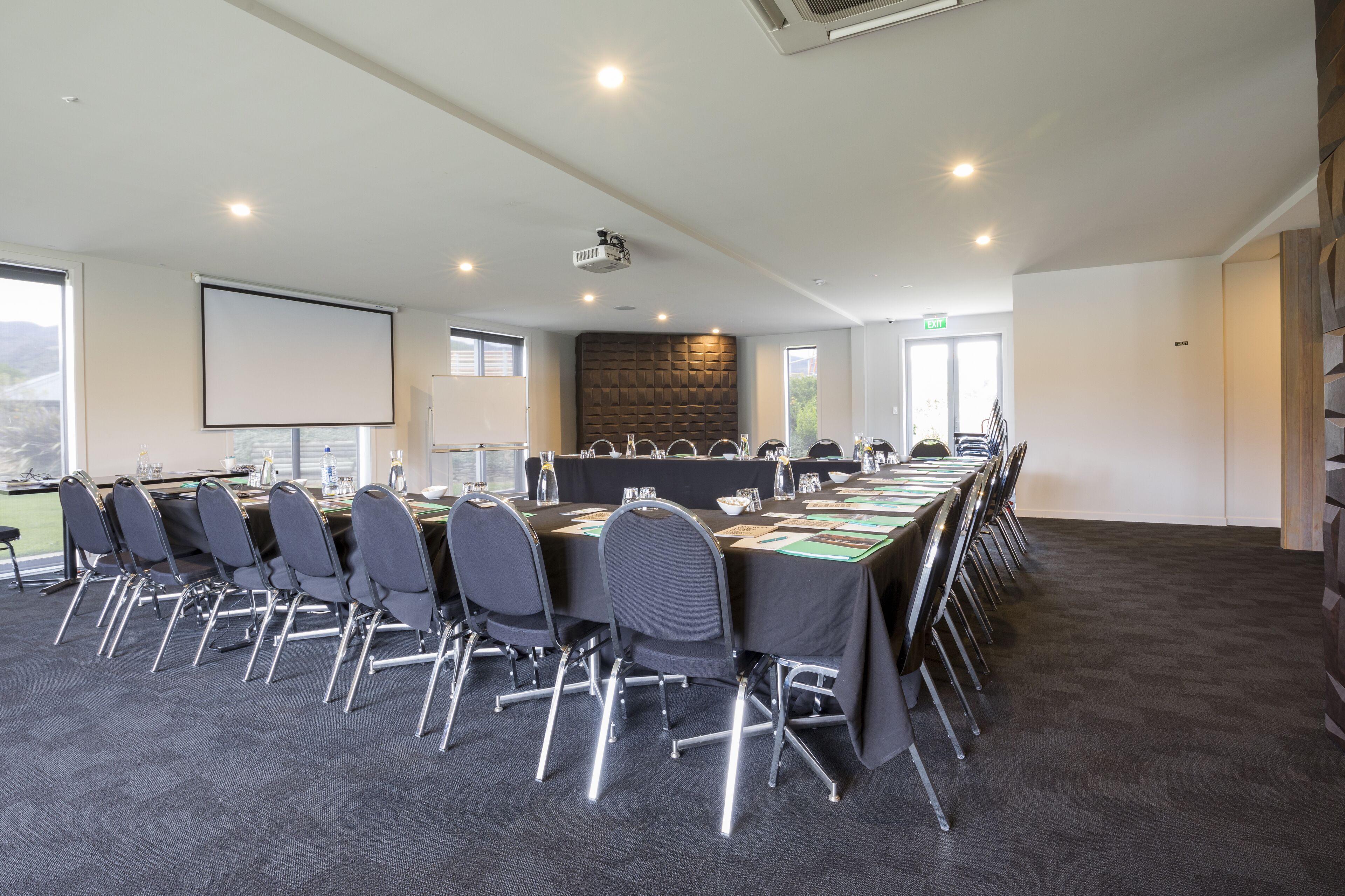 A conference room with a long table, chairs, a projector screen, and a stone wall.