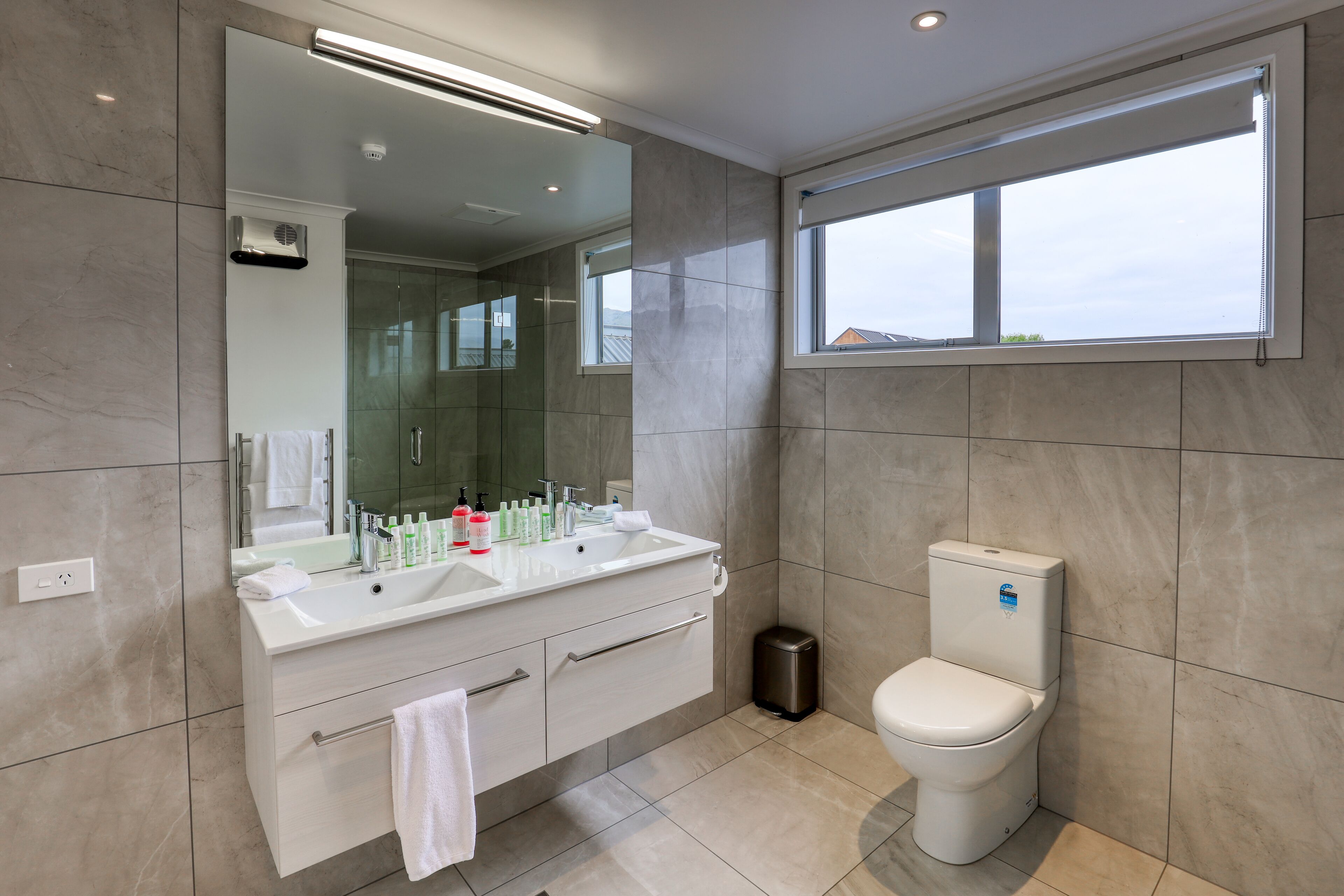 Un bagno moderno con un ampio specchio, doppio lavabo, WC e finestra che lascia entrare luce naturale.